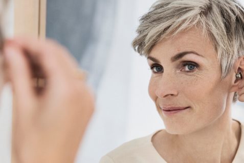 An Aged Woman Watching Her While Wearing Hearing Aid Infront Of The Mirror.