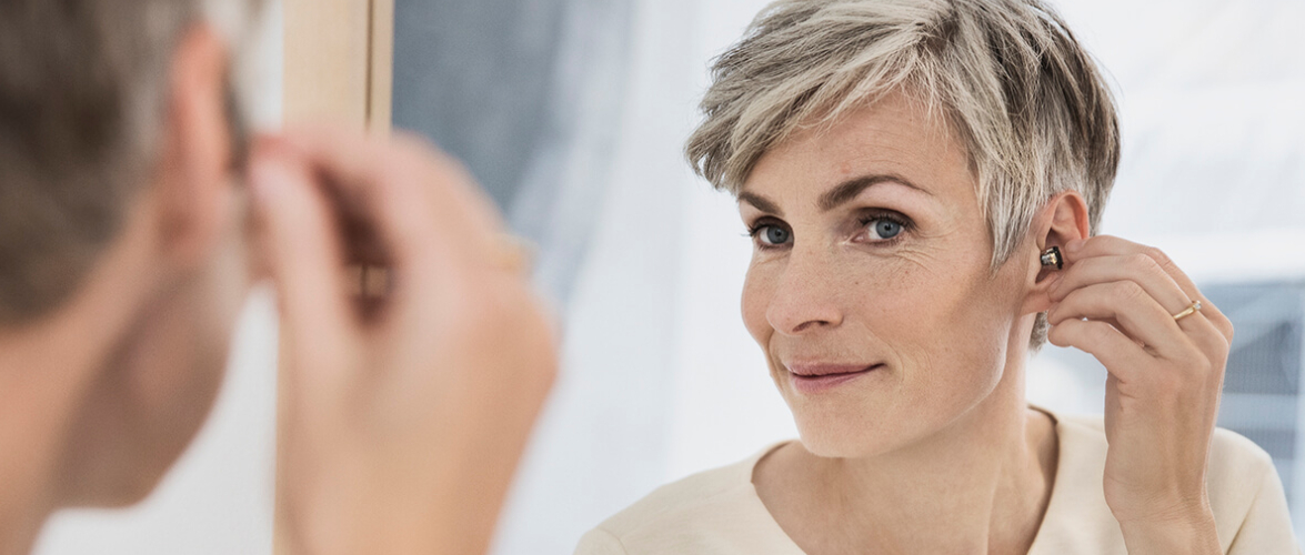 An Aged Woman Watching Her While Wearing Hearing Aid Infront Of The Mirror.